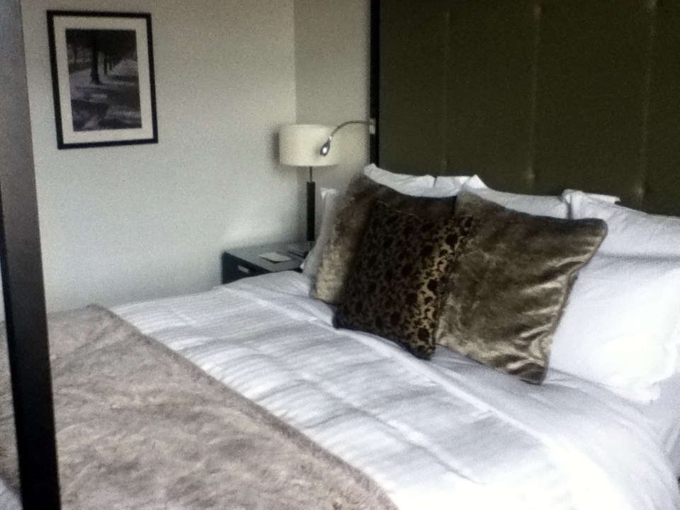 London hotel bed with white linen, decorative cushions, a bedside lamp and framed wall art.